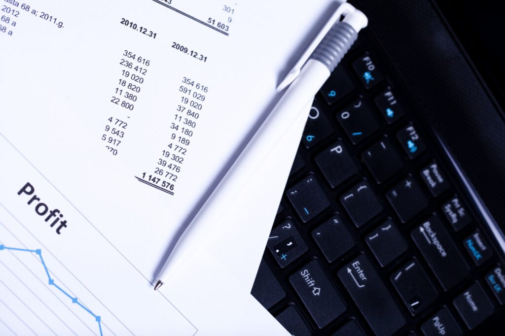 A close-up of financial documents with profit figures and charts placed over a laptop keyboard. A white pen rests on the paper, suggesting analysis or review of financial data.