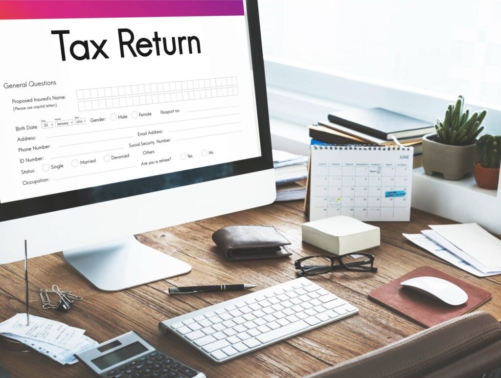 A workspace featuring a computer displaying a "Tax Return" form on the screen, surrounded by various desk items, including a keyboard, mouse, calculator, glasses, receipts, a calendar, and a few potted plants by the window. The desk has a neat yet active appearance, suggesting someone is working on their taxes.